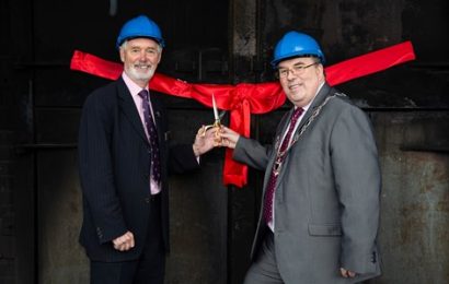 Chairman Colin Dudley and Councillor Ross standing in blue hard hats holding scissors in front of a red ribbon and firehouse doors