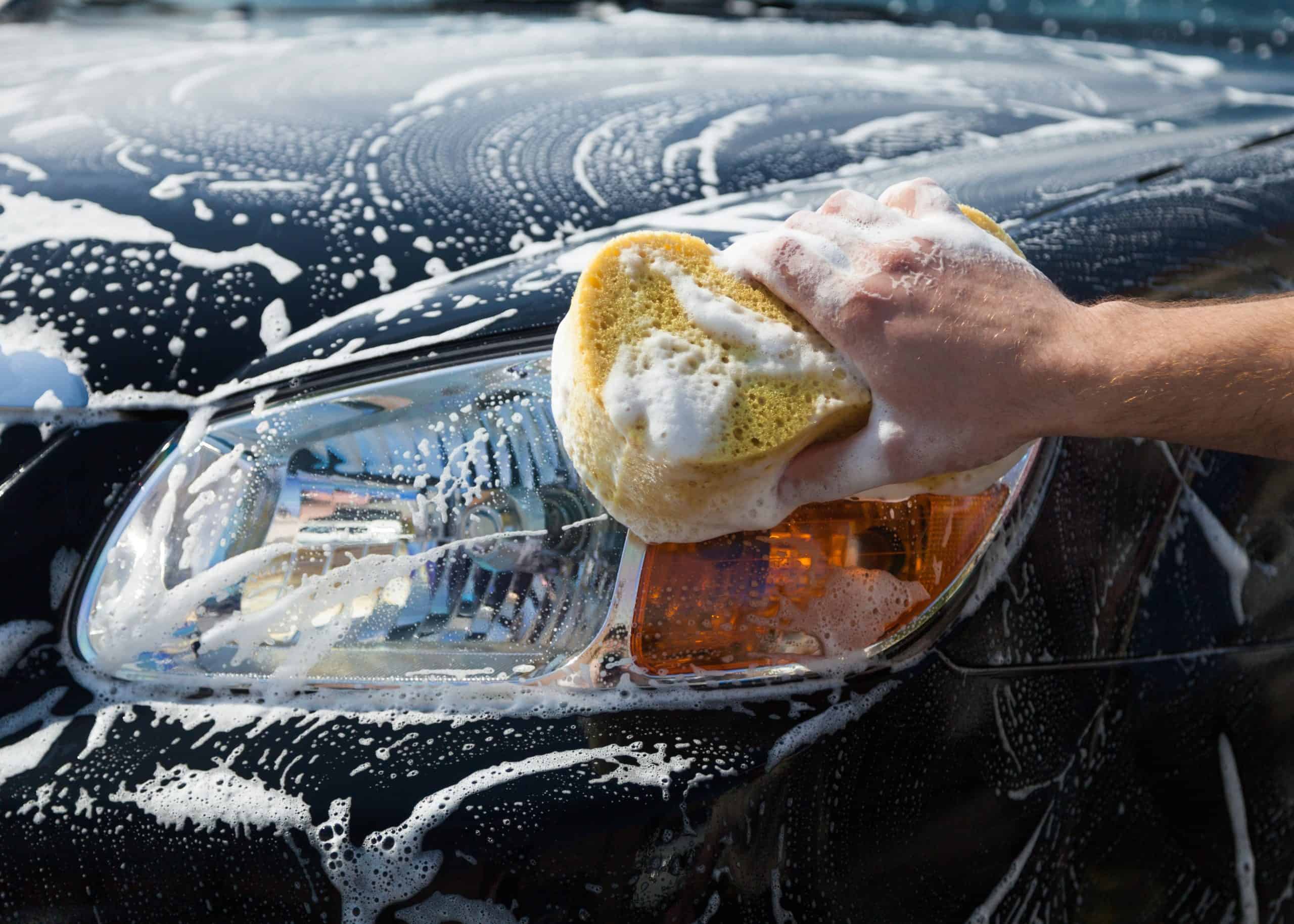 A hand holding a soapy sponge cleaning a car headlight